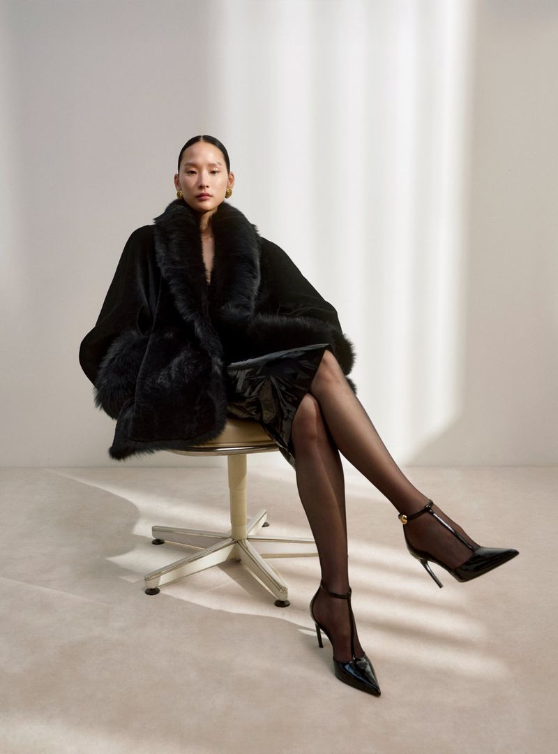 An Asian woman poses while sitting on a chair, wearing a black fur coat, a shiny dress, and black high heels against a neutral backdrop.