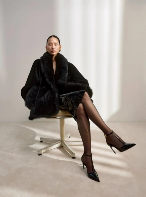 An Asian woman poses while sitting on a chair, wearing a black fur coat, a shiny dress, and black high heels against a neutral backdrop.