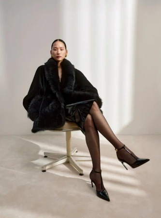 An Asian woman poses while sitting on a chair, wearing a black fur coat, a shiny dress, and black high heels against a neutral backdrop.