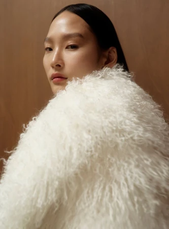 Model Yoonmi Sun wearing a high-collared white curly shearling coat, looking over her shoulder against a warm brown backdrop.
