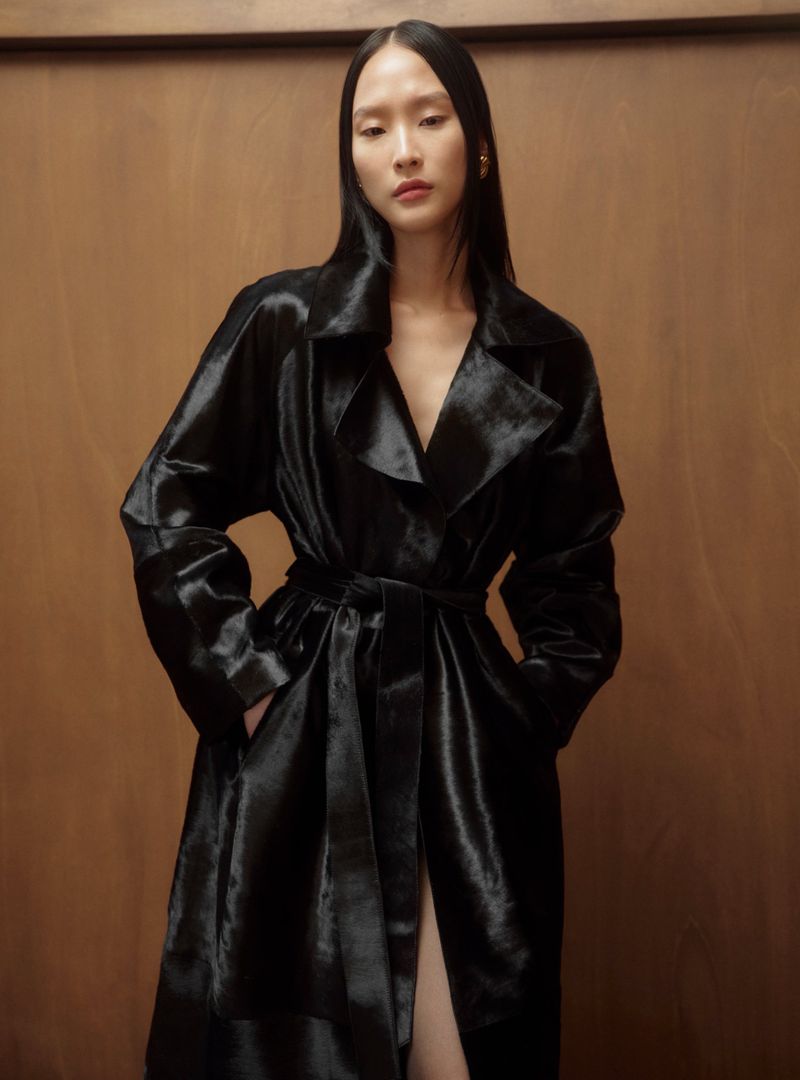 An Asian woman with long dark hair poses in a shiny black belted trench coat against a wood-panelled wall, looking directly at the camera.
