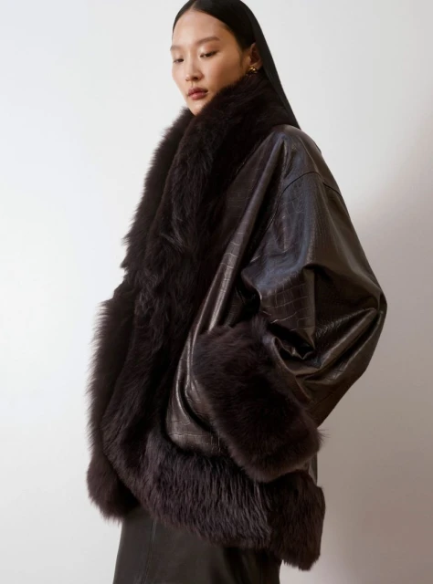 Brunette Asian model showcases a dark brown, textured, oversized jacket trimmed with dark fur against a plain background in a studio shot.