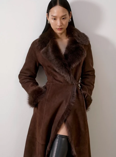 Elegant woman poses indoors, wearing a brown fur-trimmed suede wrap coat with a tie belt and black knee-high boots against a neutral backdrop.