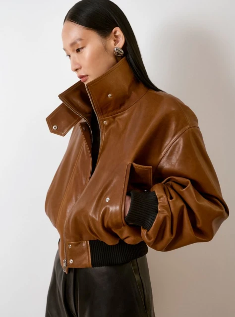 An Asian female model showcases a brown leather bomber jacket with a high collar in a fashion portrait, posing indoors on a neutral background.