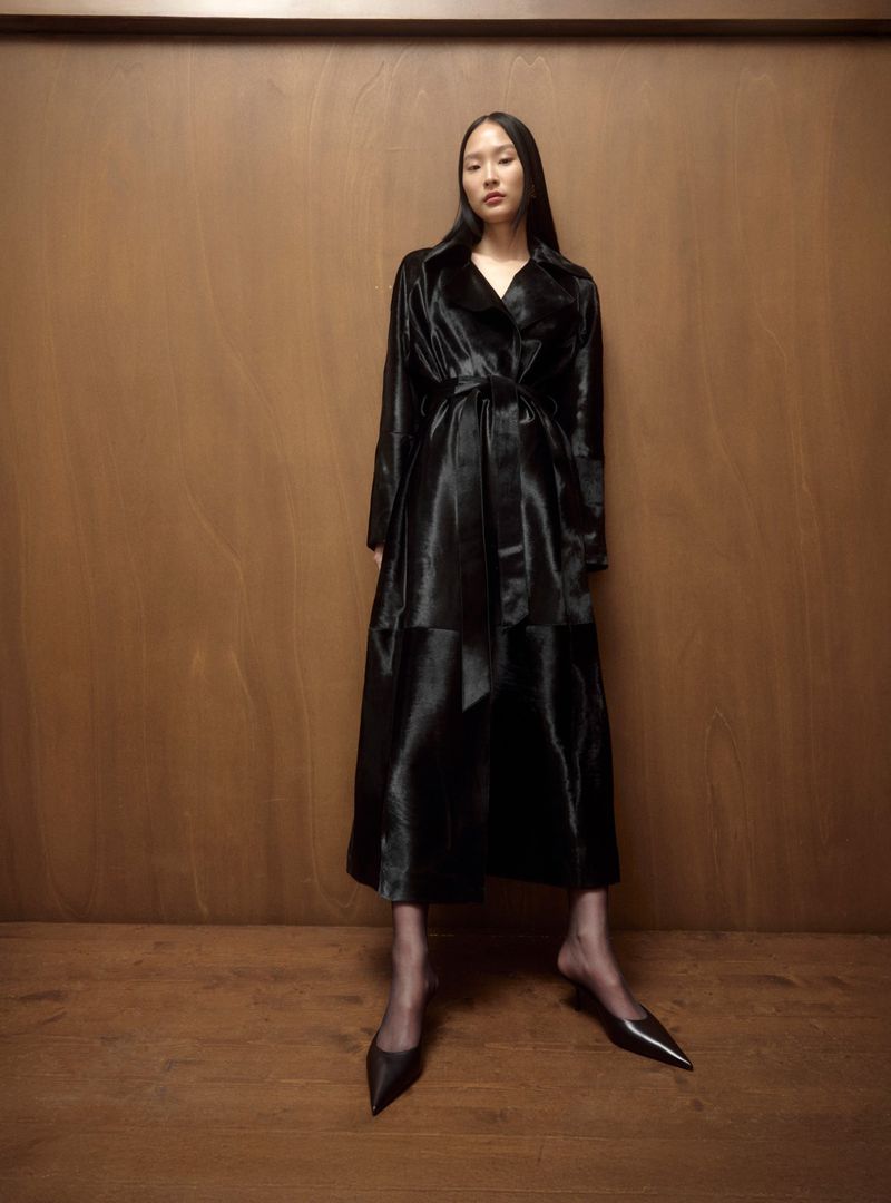 An Asian woman poses indoors wearing a long, shiny black coat with a tie belt, paired with black heels against a brown wood background.