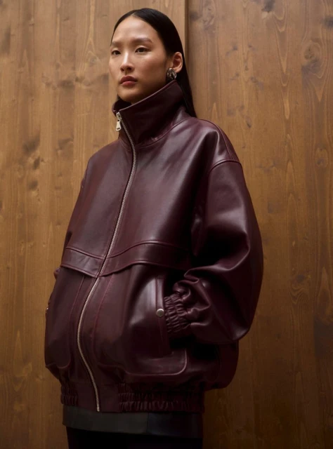 Portrait of an Asian woman wearing a burgundy leather jacket with a high collar against a light brown wood panel background.