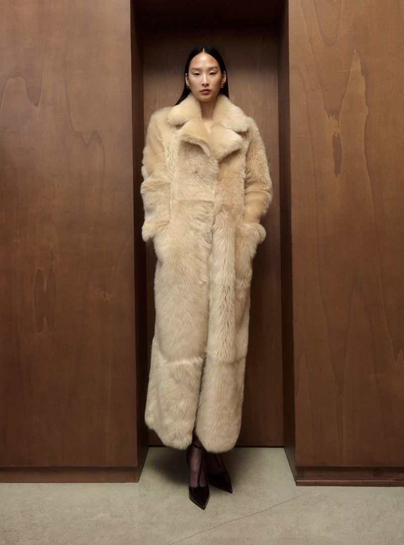 An Asian woman stands in a warm, neutral-toned room, modeling a full-length, beige shearling coat with pointed black shoes, striking an editorial pose.