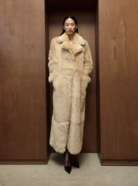 An Asian woman stands in a warm, neutral-toned room, modeling a full-length, beige shearling coat with pointed black shoes, striking an editorial pose.