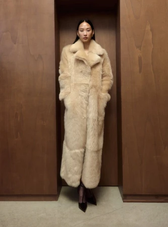 An Asian woman stands in a warm, neutral-toned room, modeling a full-length, beige shearling coat with pointed black shoes, striking an editorial pose.