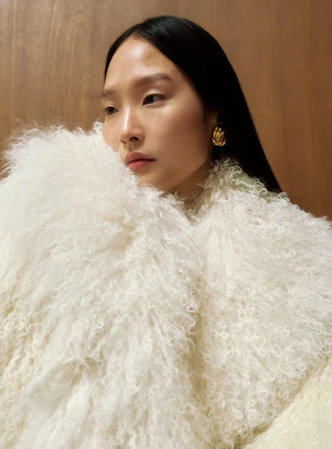 Close-up of an Asian woman with dark hair and gold earrings, wearing a shaggy white coat against a neutral-toned wooden background.