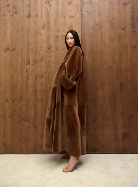 A woman in a brown coat poses in front of a wood-paneled wall wearing translucent heeled shoes in a studio shot.