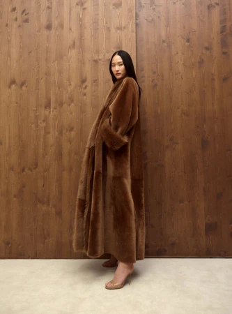 A woman in a brown coat poses in front of a wood-paneled wall wearing translucent heeled shoes in a studio shot.