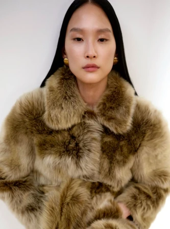 Model Yoonmi Sun wearing a plush collared faux fur coat and gold earrings against a bright white studio backdrop.