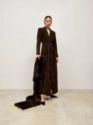 Model wearing a dark brown belted coat holding a long fur stole against a neutral studio background.