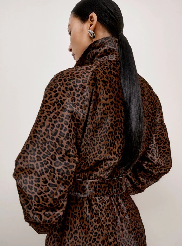 Model with long dark ponytail wearing a leopard print belted coat against a plain background
