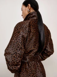 Model with long dark ponytail wearing a leopard print belted coat against a plain background
