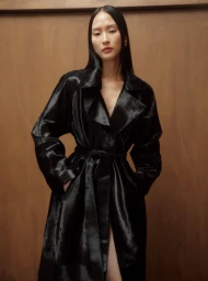 Model wearing a glossy black belted coat against a warm brown wooden wall interior background