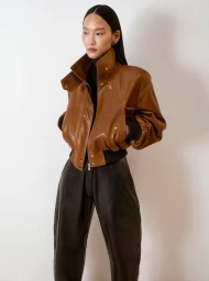 Model Yoonmi Sun wears a caramel brown leather bomber jacket with a high structured collar and black leather trousers against a plain studio backdrop.
