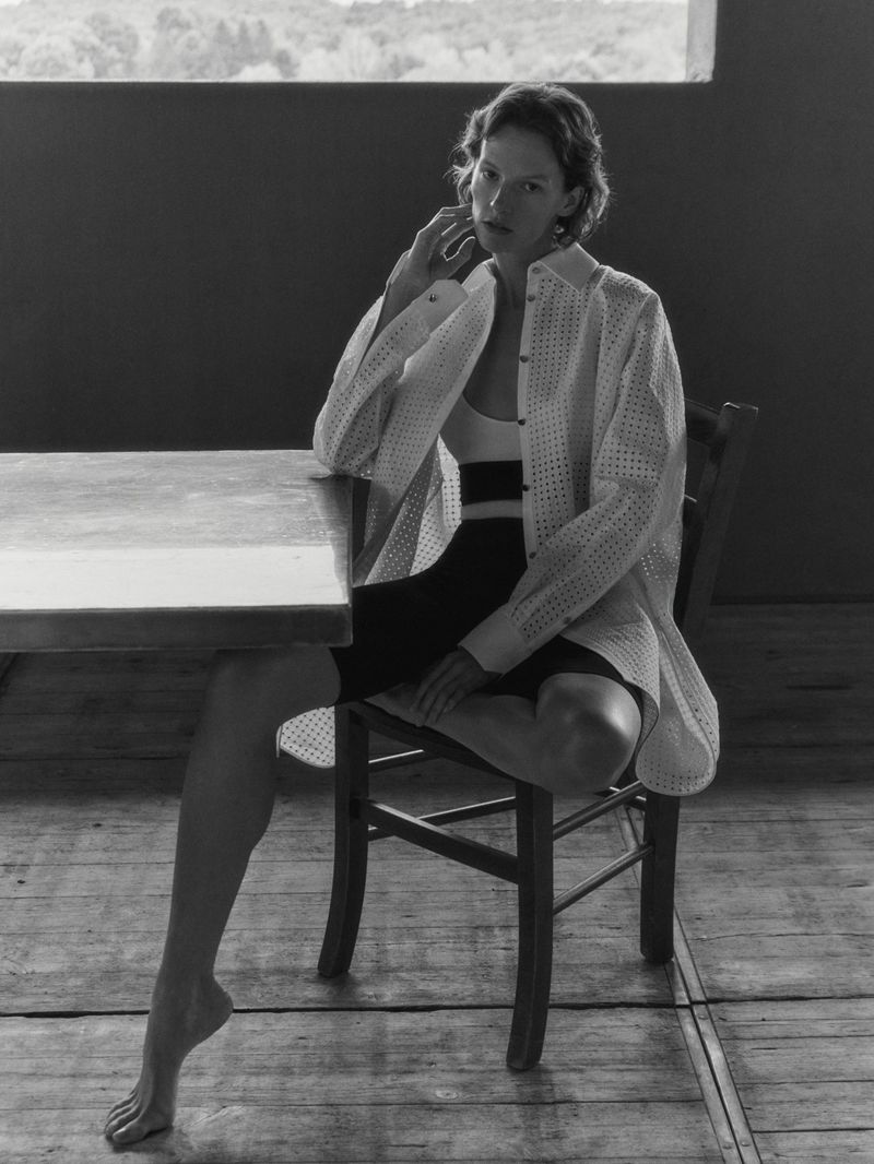 Monochrome portrait of a woman in an unbuttoned, patterned white blouse sitting at a table and chair inside a simple room.