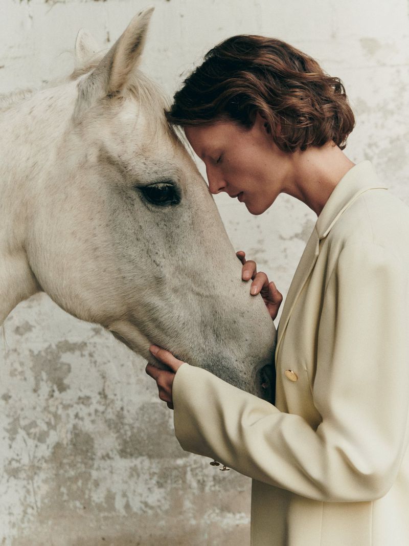 Woman in cream jacket embraces a white horse in a close-up shot with a neutral background, focusing on connection and natural tones.