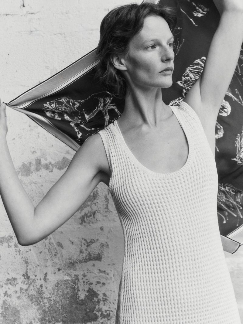 Woman in white dress poses with patterned scarf against textured wall in black and white studio shot, looking away with casual pose.
