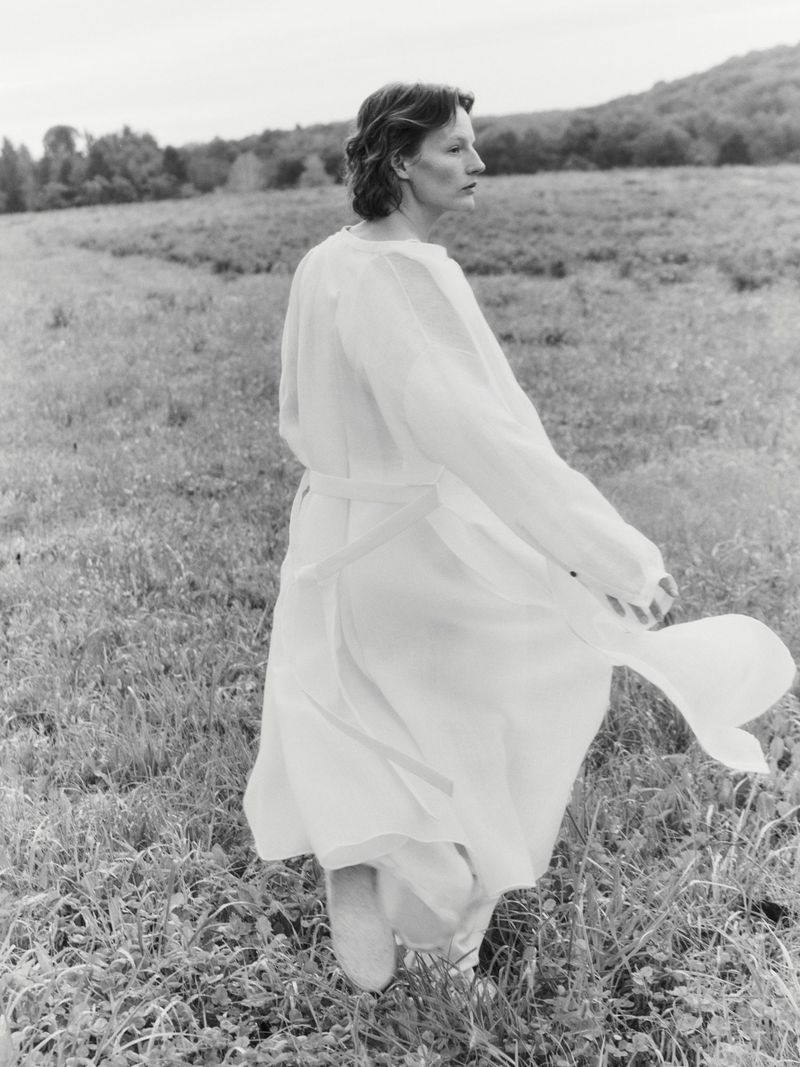 Monochrome portrait of a woman in a flowing white dress standing in a grassy field with hills and trees in the background, outdoor shot.