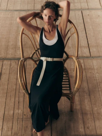 A woman with short hair in a black dress with white trim poses while seated in a wicker chair in a minimalist indoor setting.