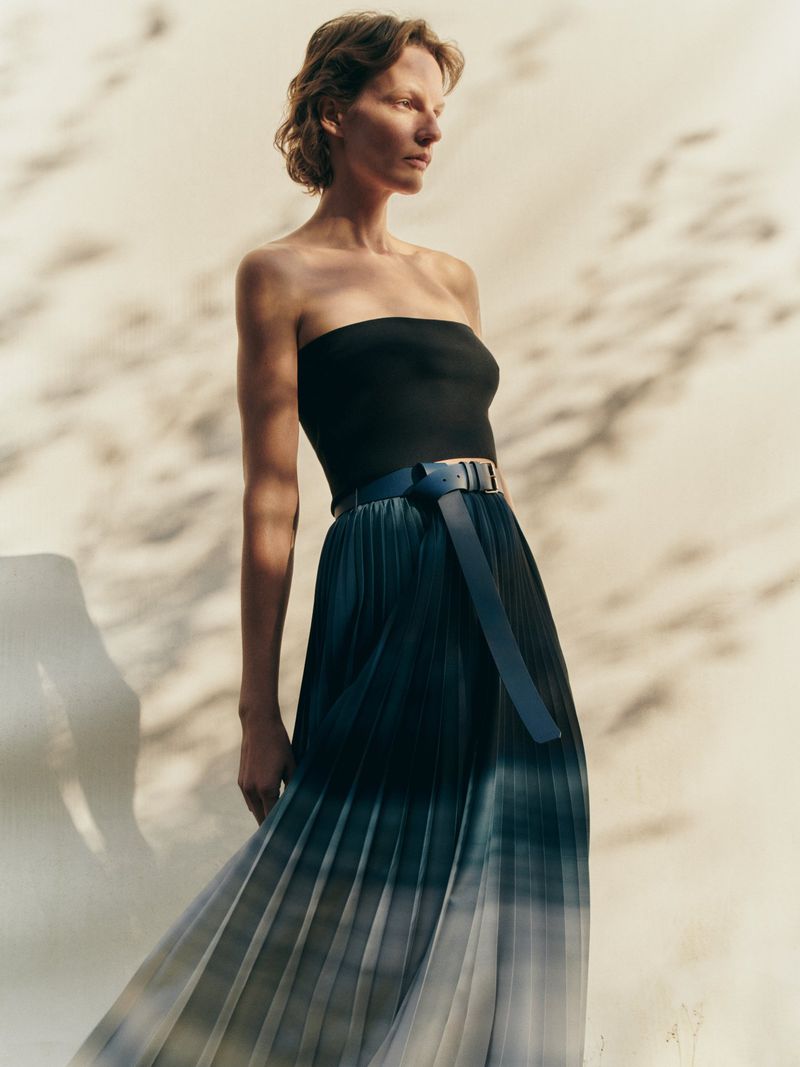 Model wearing a black bandeau top and pleated ombré skirt against a sandy textured wall with soft shadows.