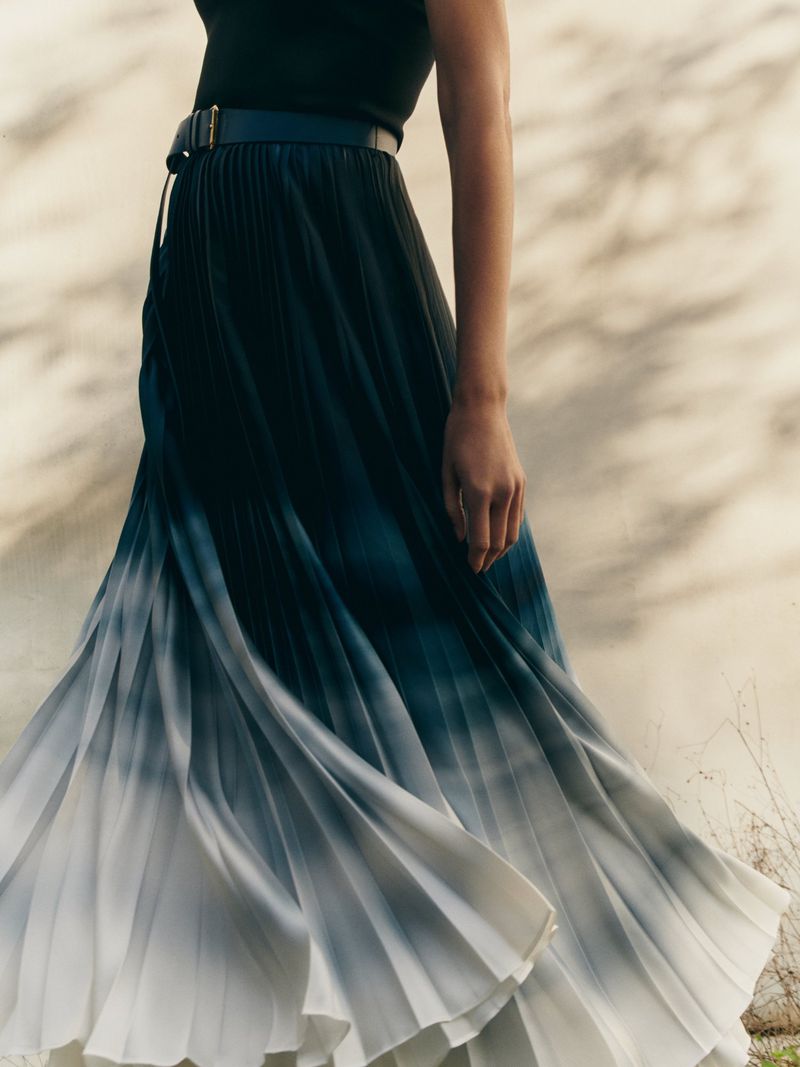 A woman models a flowing pleated midi skirt with an ombre effect transitioning from dark blue to white against a neutral backdrop.