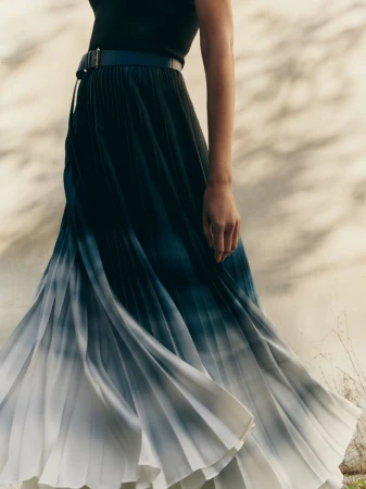 A woman models a flowing pleated midi skirt with an ombre effect transitioning from dark blue to white against a neutral backdrop.