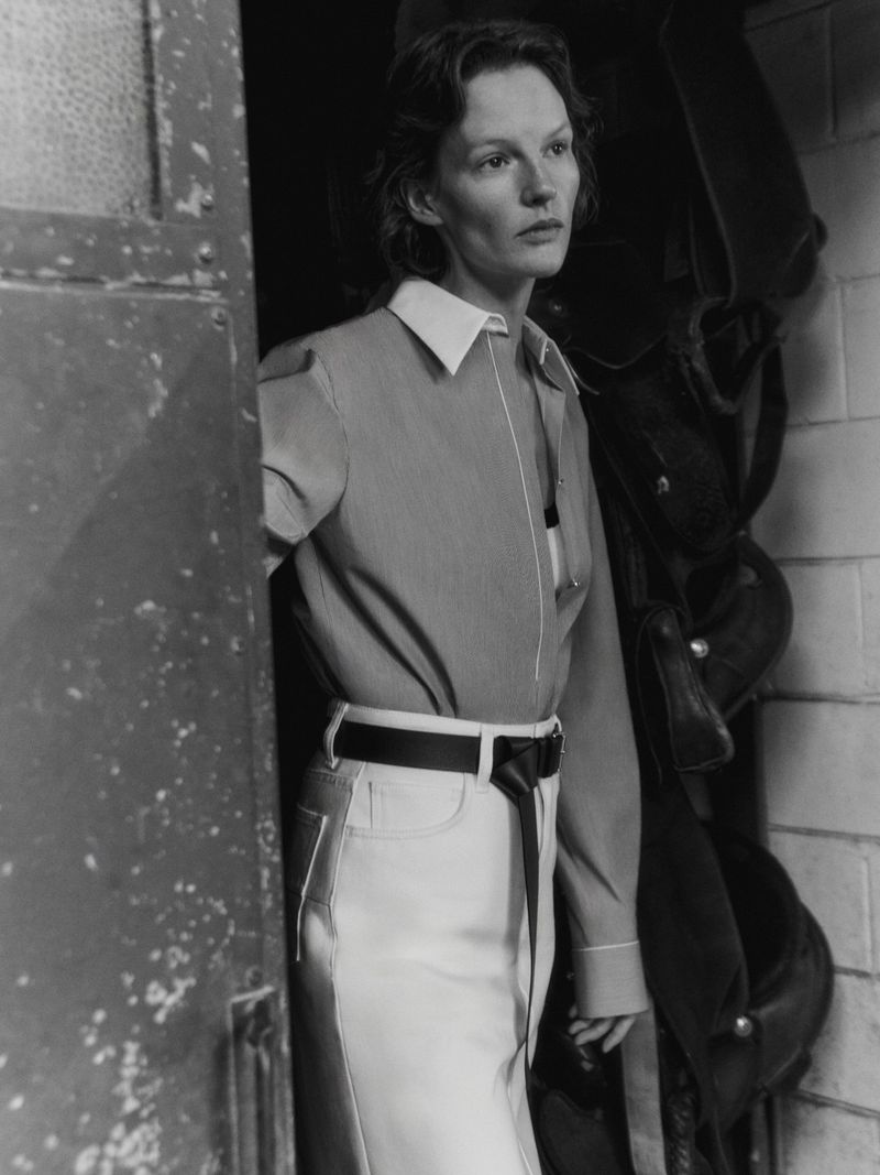 Monochrome portrait of a stylish adult woman in a collared shirt and white skirt, posing near a weathered doorway with riding gear.