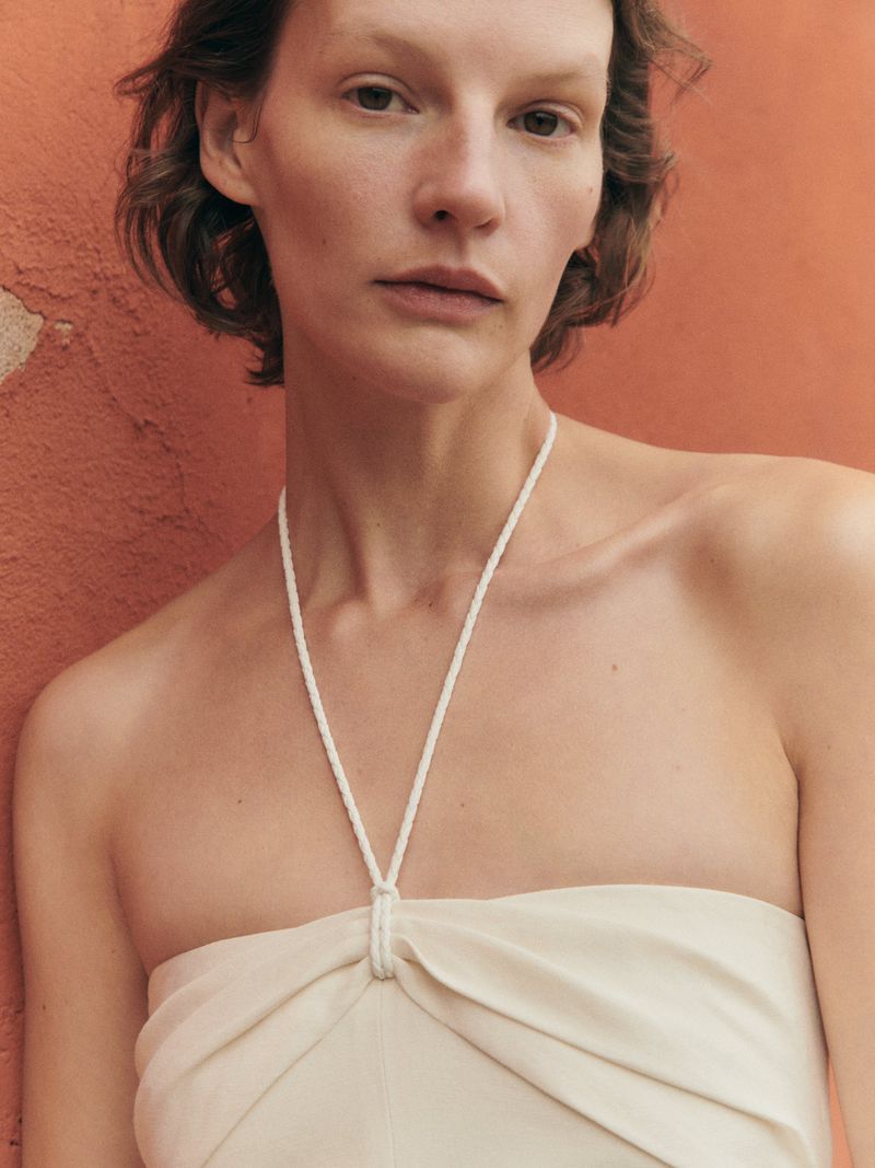 Close-up of an adult woman in a light-colored halter dress against a textured, solid orange background in a fashion-style composition.