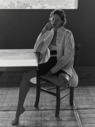 Sara Blomqvist seated on a wooden chair by a table wearing an unbuttoned white perforated shirt for Giada.