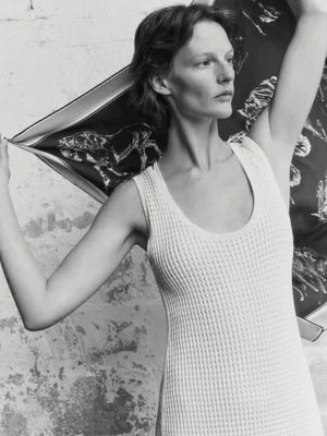 Model wearing a textured sleeveless knit dress holding a patterned scarf against a weathered textured wall background