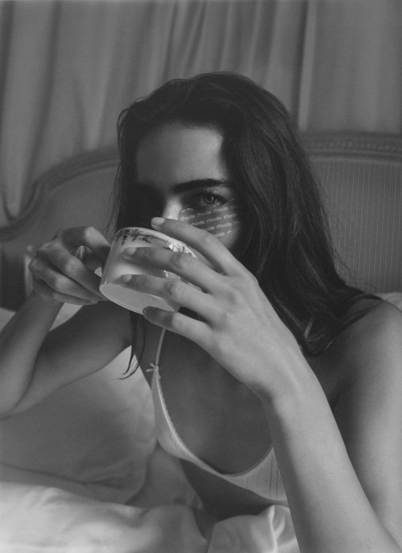 A woman is photographed indoors, holding a teacup while lying in bed. The image is black and white with muted tones.