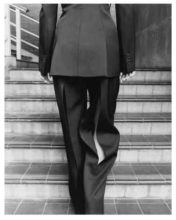 Monochrome studio shot from behind of a person on stairs wearing a suit with split-seam trousers, minimalist fashion.