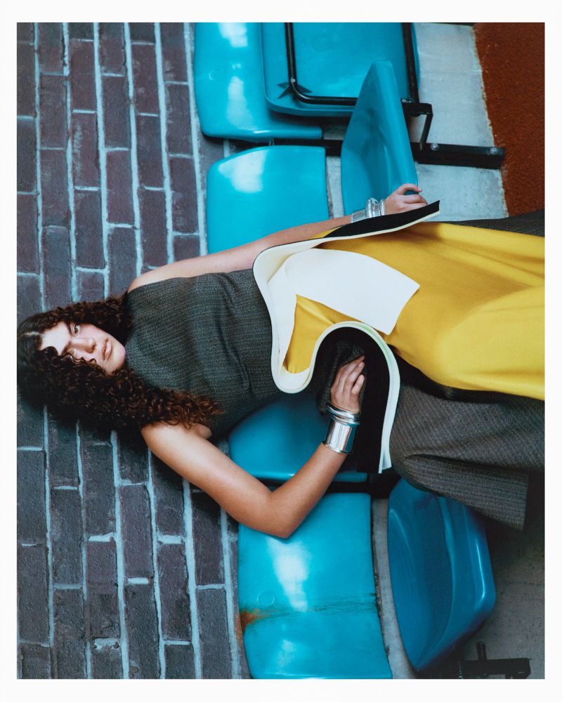 Fashion shot featuring a curly-haired woman in a unique dress, reclining amongst blue chairs against a brick wall backdrop, creating a surreal scene.