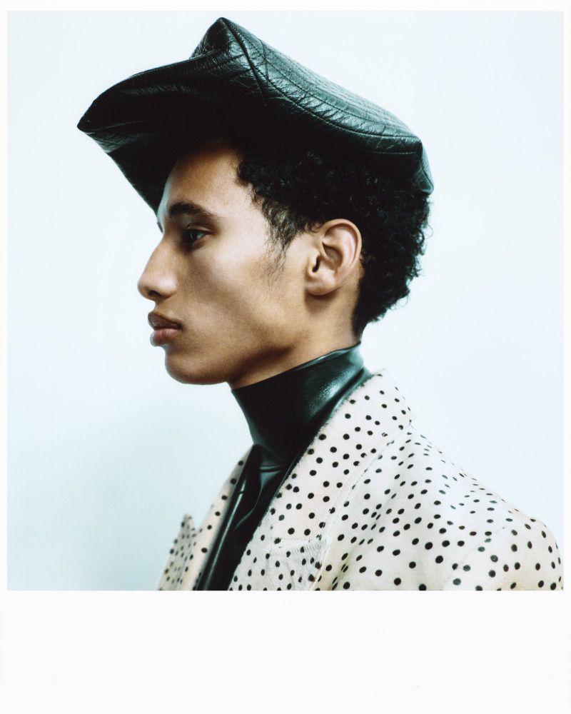Portrait of adult male in black leather-like hat and polka-dot coat against plain backdrop in studio lighting. Fashion editorial shot.