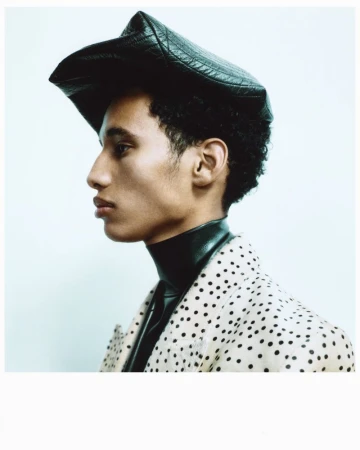 Portrait of adult male in black leather-like hat and polka-dot coat against plain backdrop in studio lighting. Fashion editorial shot.