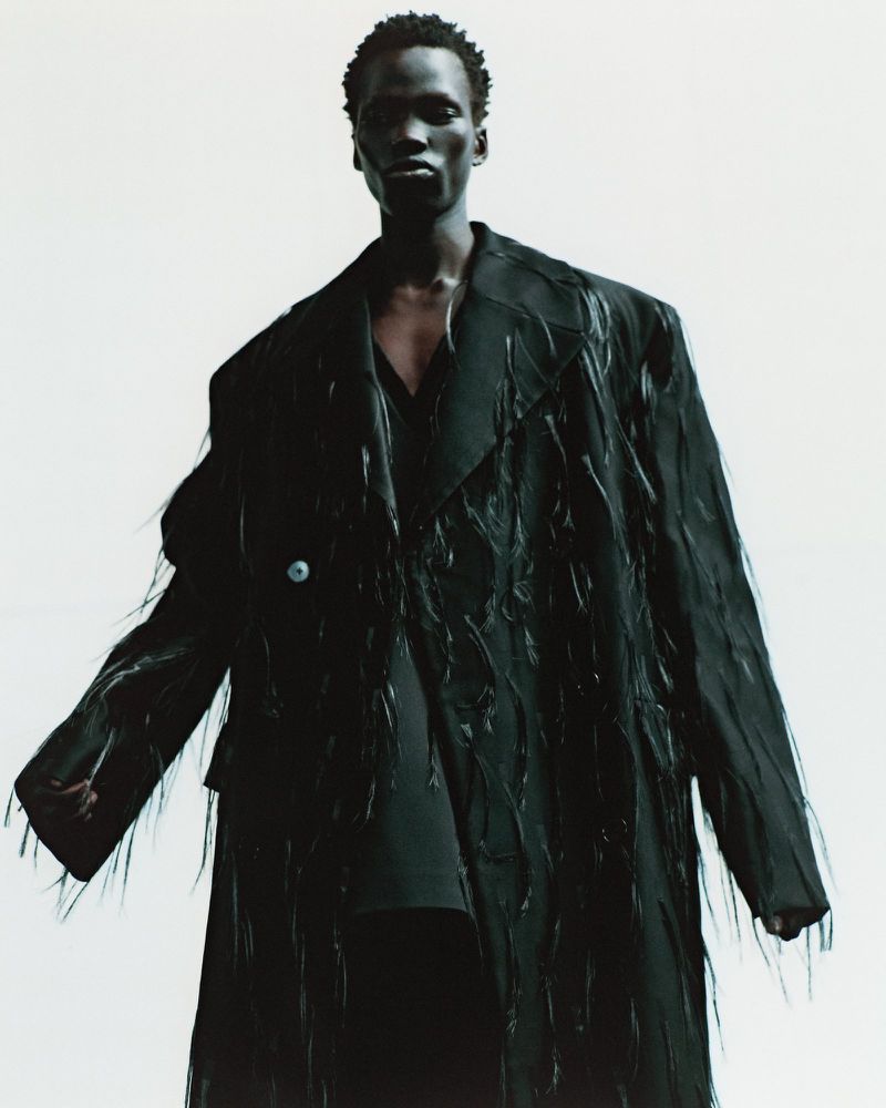 Portrait of African male model in a dark, textured overcoat against a bright white background in a fashion photography setting.