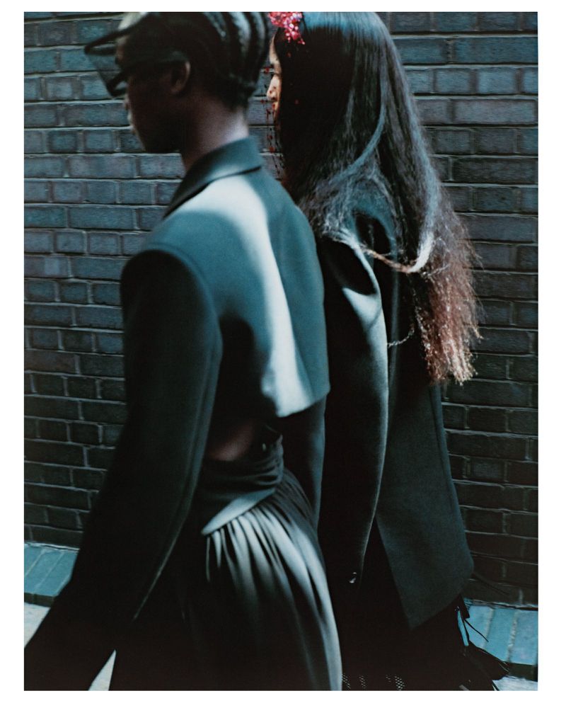 Two women in dark blazers walk closely together along a brick wall. One has long hair, and the other is wearing a cap.