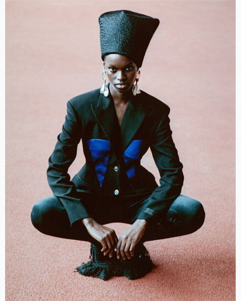 An African American fashion model squats in a studio, wearing a black jacket with blue bodice, black jeans, a tall hat, and big earrings.