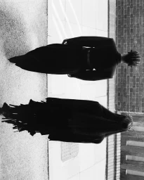 Two models seen from behind wearing structured black garments standing on a sunny paved street next to a brick wall in black and white.