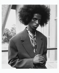 Monochrome portrait of a model with voluminous afro hair wearing an oversized blazer and patterned shirt outdoors.