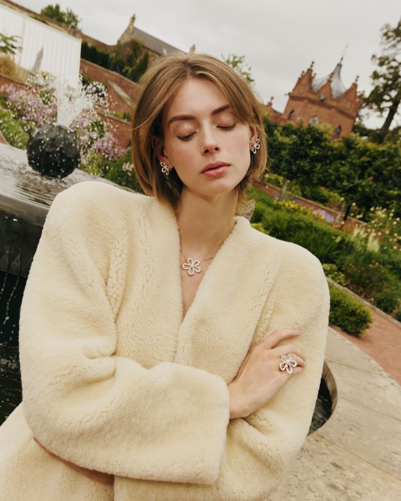 Model Lauren de Graaf in a cream fleece coat wearing floral diamond jewelry in an outdoor estate garden