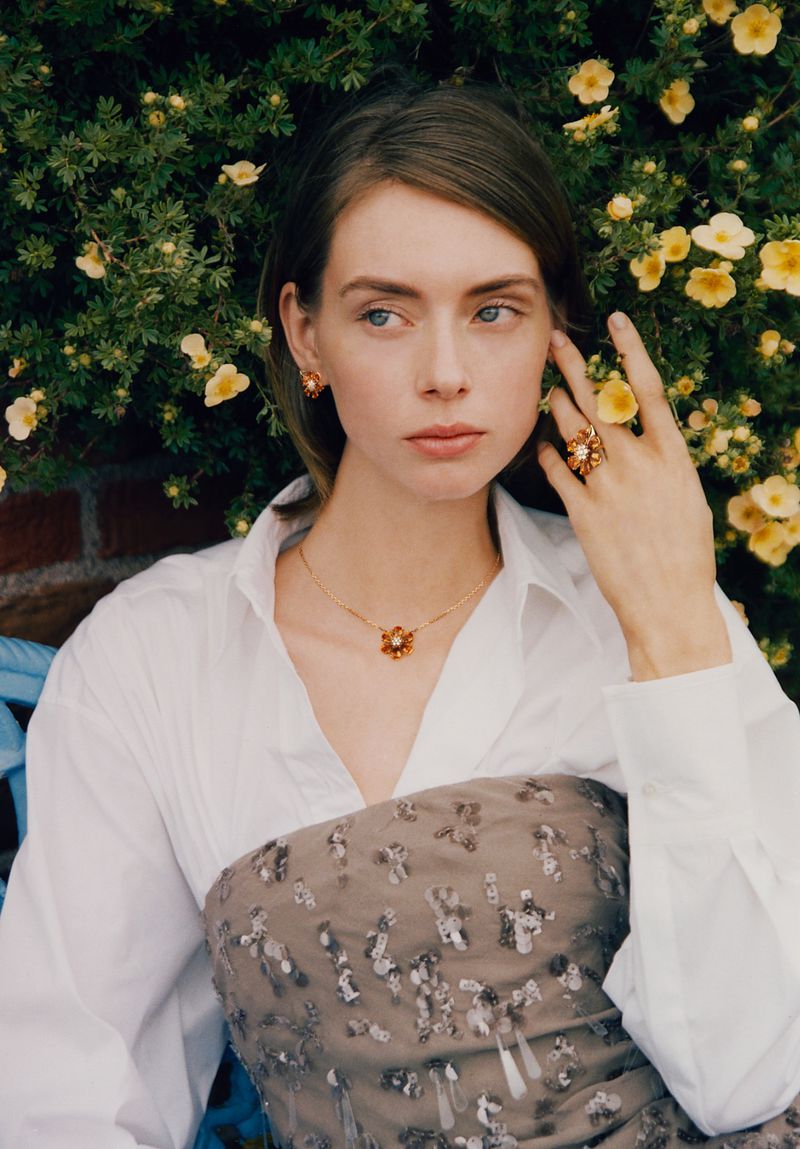 Portrait of a fair-skinned young woman wearing gold floral jewelry, including earrings, necklace, and ring, posed in front of a flowering bush.