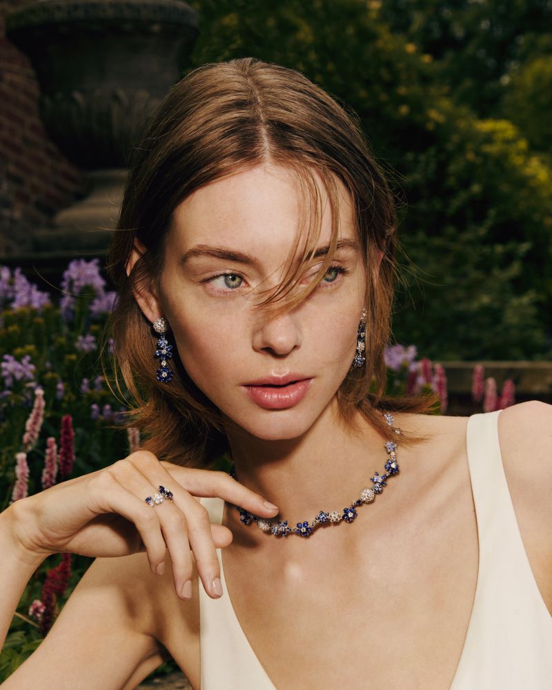 Close-up of a young woman wearing a floral jewelry set including a necklace, earrings, and a ring posing in a garden setting.