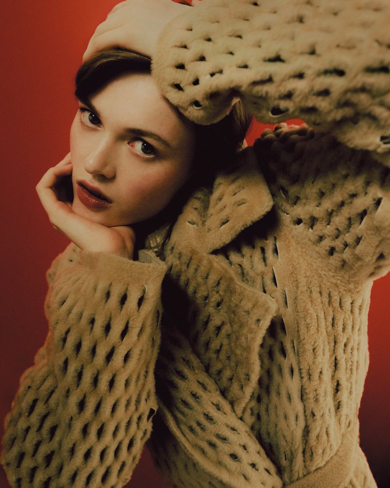 Portrait of a fashionable woman with fair skin wearing a fuzzy beige coat and matching beret posing against a red background.