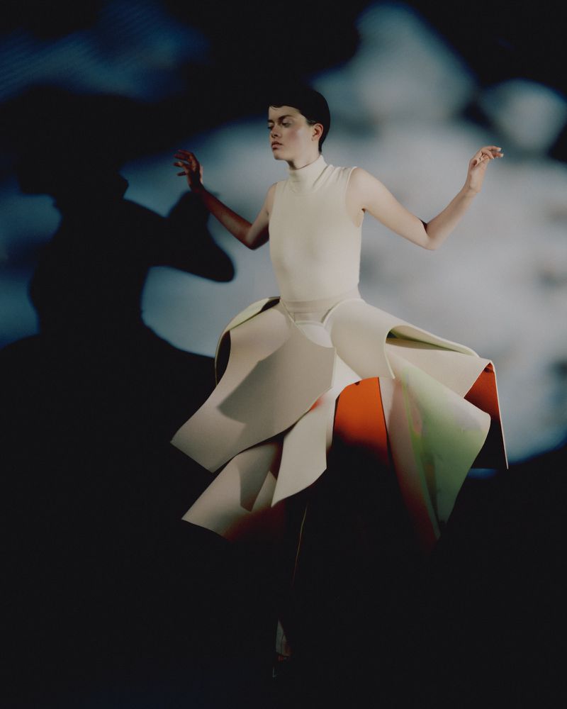 Young woman strikes a pose in a cream-colored dress with an origami-like skirt against a cloudy backdrop in a studio setting. 