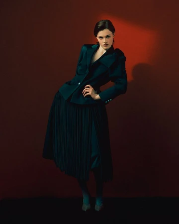 Model posed in a dark teal structured blazer and matching pleated midi skirt against a deep crimson studio backdrop.